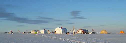 The camp at around lunch time (midnight) as seen from the north. From left to right: Fuel drums and cargo, generator tent, ice core freezer, drill tent (large white construction), kitchen tent, food storage, medical tent, and sleeping tent. The pole in the front is the weather station.